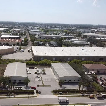 Aerial view of an industrial area with large warehouses, office buildings, parked vehicles, and surrounding roads on a clear day.