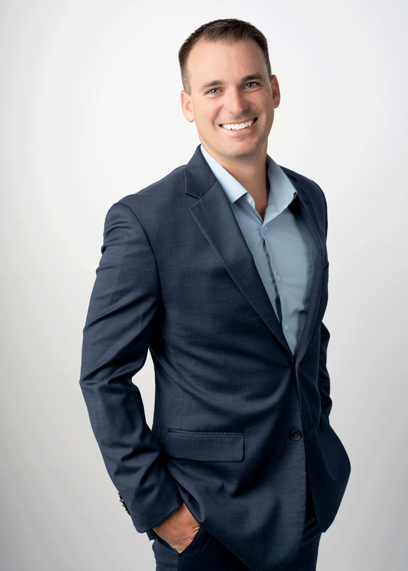 Man in a blue suit with a light blue shirt stands against a plain light background, smiling with one hand in his pocket.