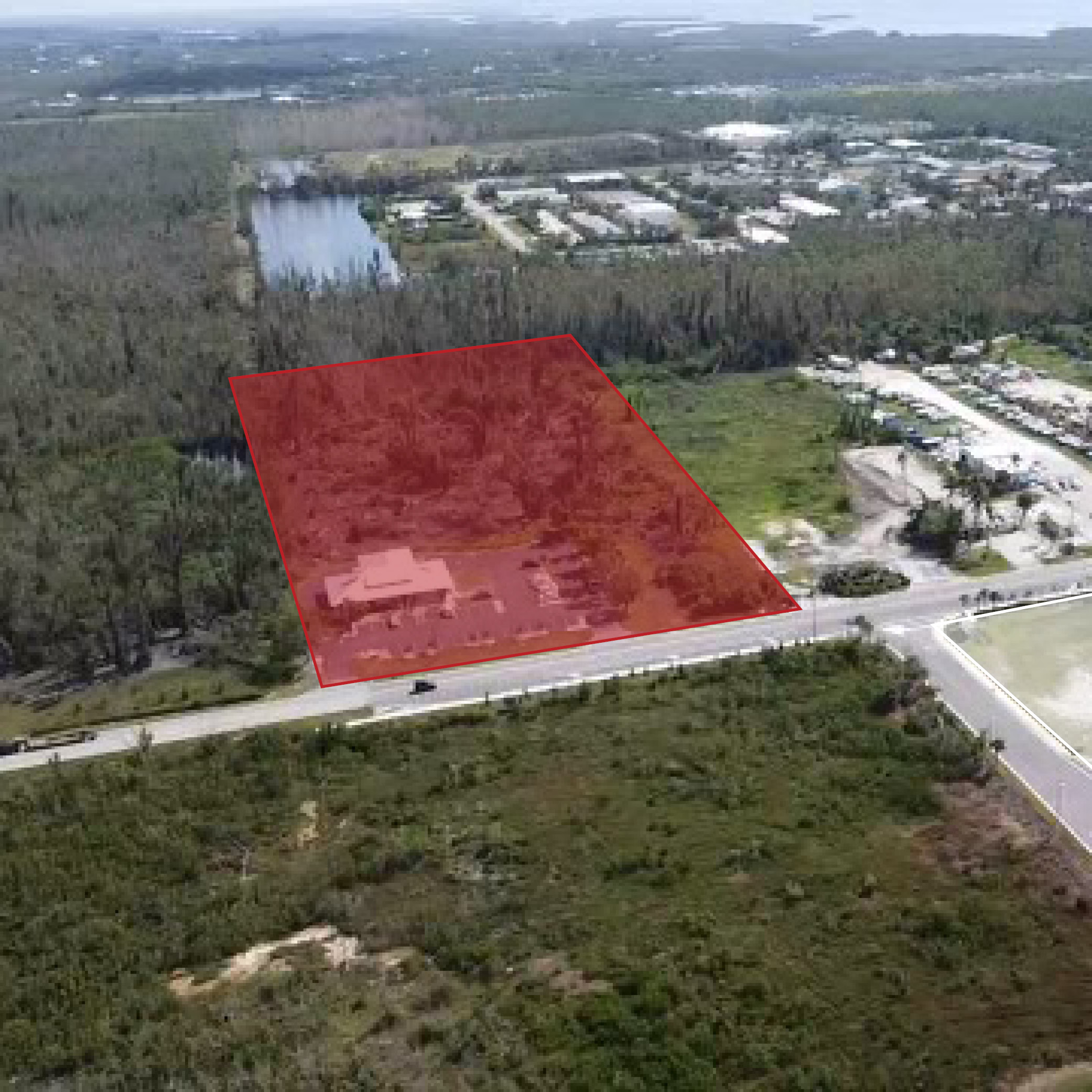 Aerial view of a plot of land near a road, highlighted in red, with buildings and trees in the background.