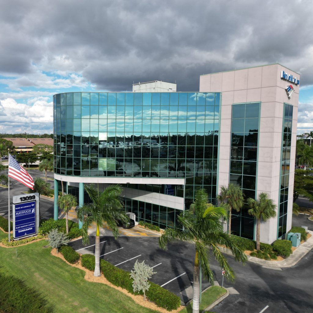 A modern glass office building with palm trees, an American flag, and a Bank of America logo, set under a cloudy sky.