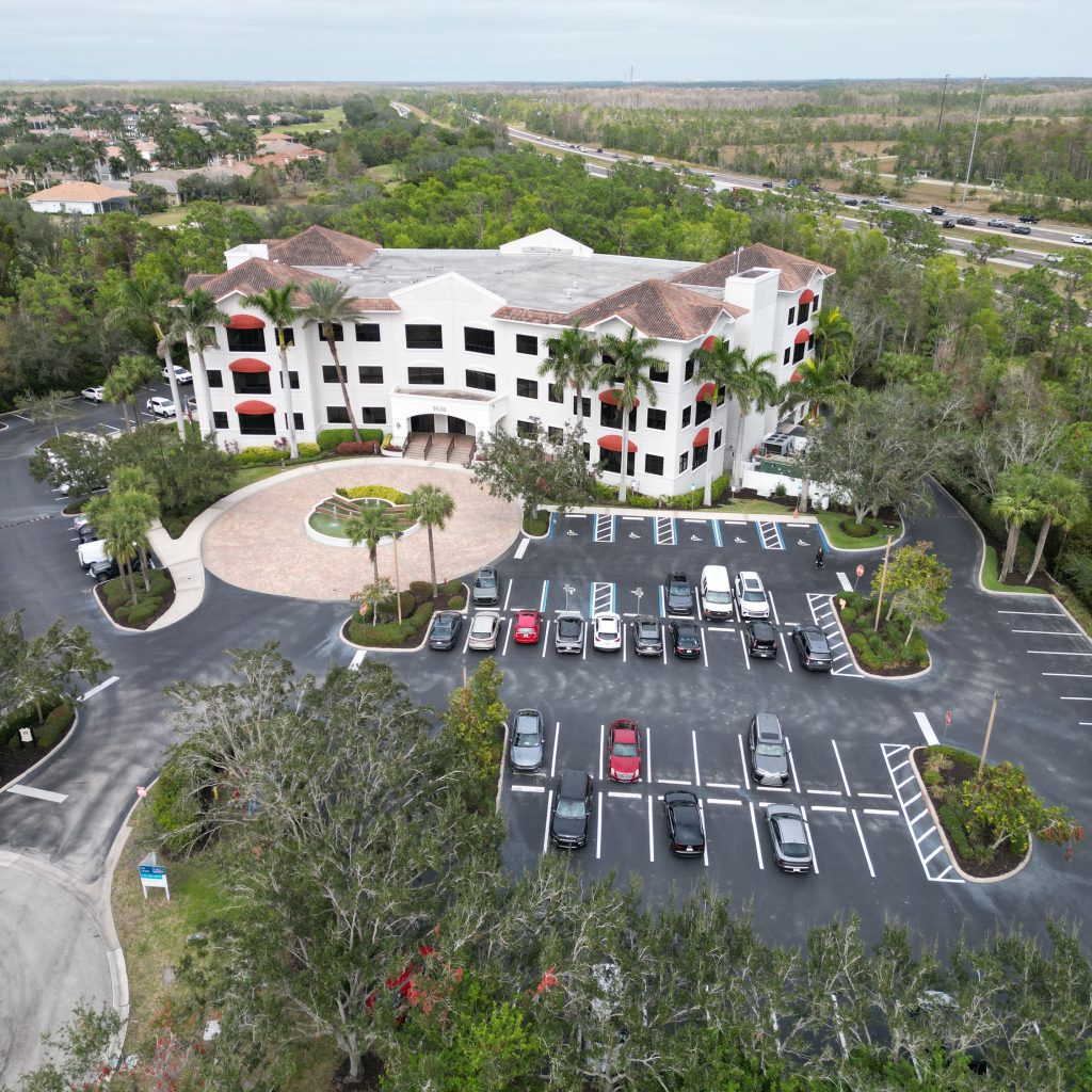 Aerial view of a three-story office building with palm trees, surrounded by a parking lot with several cars and landscaped greenery.