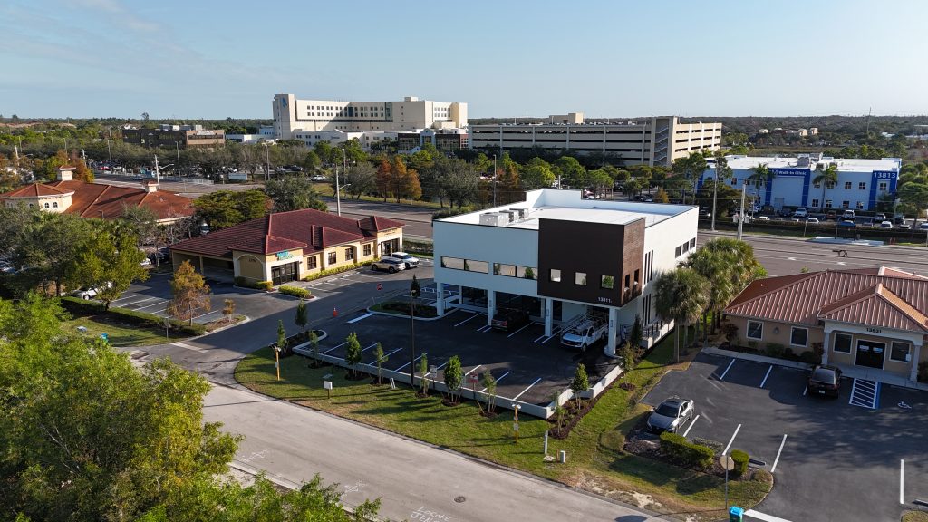 Aerial view of a small office building with a parking lot, surrounded by roads, other buildings, and trees under a clear sky.