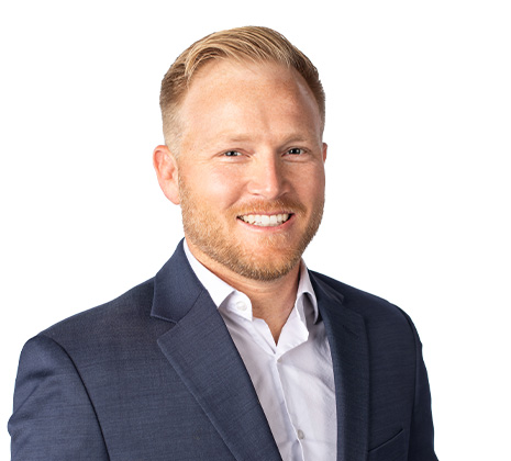 A man with short blond hair and a trimmed beard, wearing a dark suit jacket and white shirt, smiles at the camera against a plain white background.