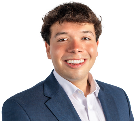 A young man with short brown hair, wearing a navy blue blazer and white shirt, smiling against a plain white background.