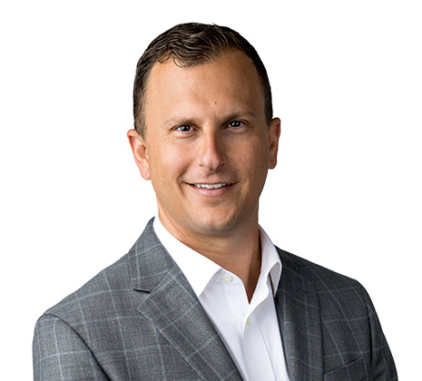A man in a gray plaid suit jacket and white shirt smiles at the camera against a plain white background.
