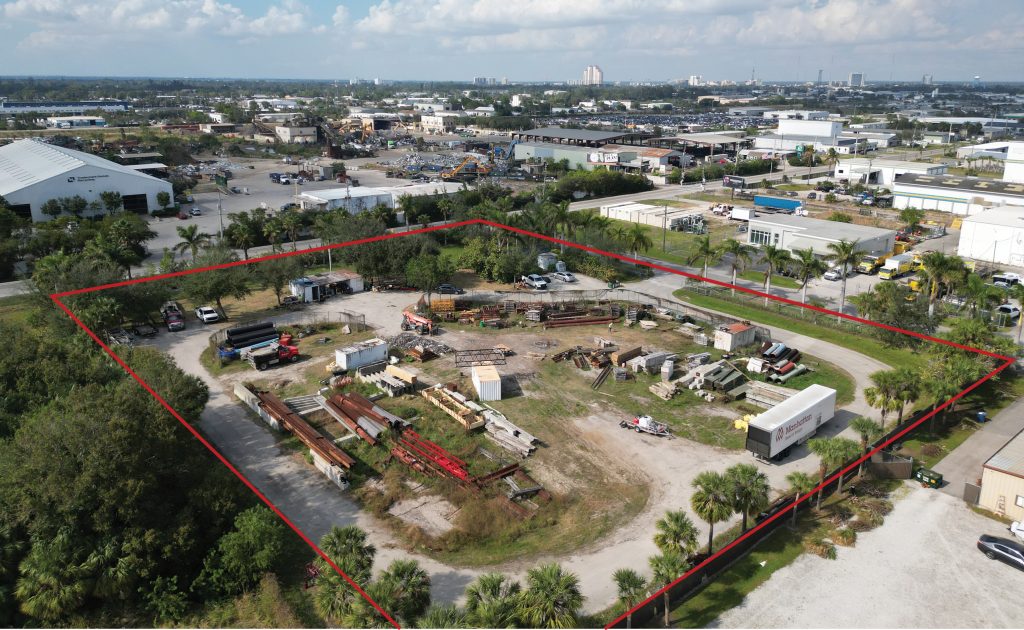 Aerial view of an industrial lot outlined in red, containing equipment, vehicles, and materials, surrounded by buildings and trees in an urban area.