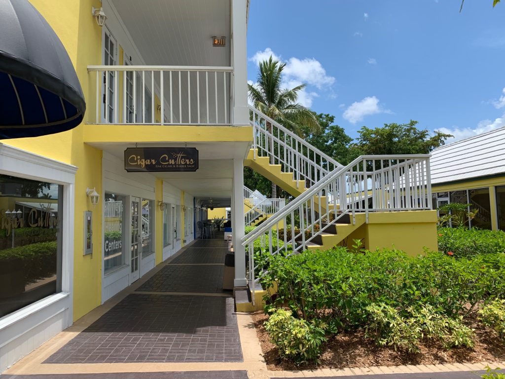 A yellow building with white railings and stairs, featuring a "Cigar Cutters" sign, lush greenery, and a partly cloudy blue sky.
