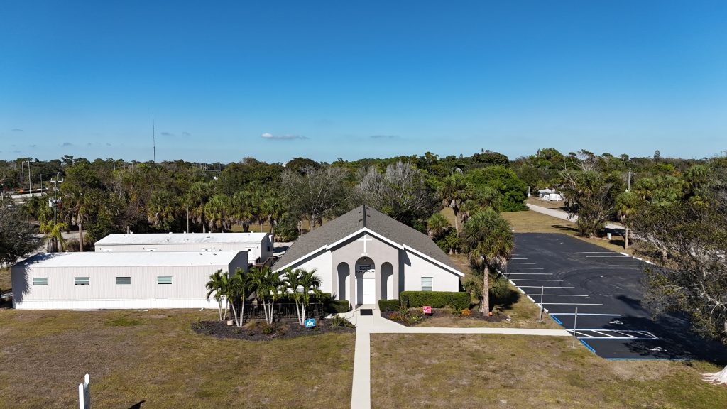 A white building with an arched entrance and adjacent annex sits amid palm trees, with a mostly empty parking lot under a clear blue sky.