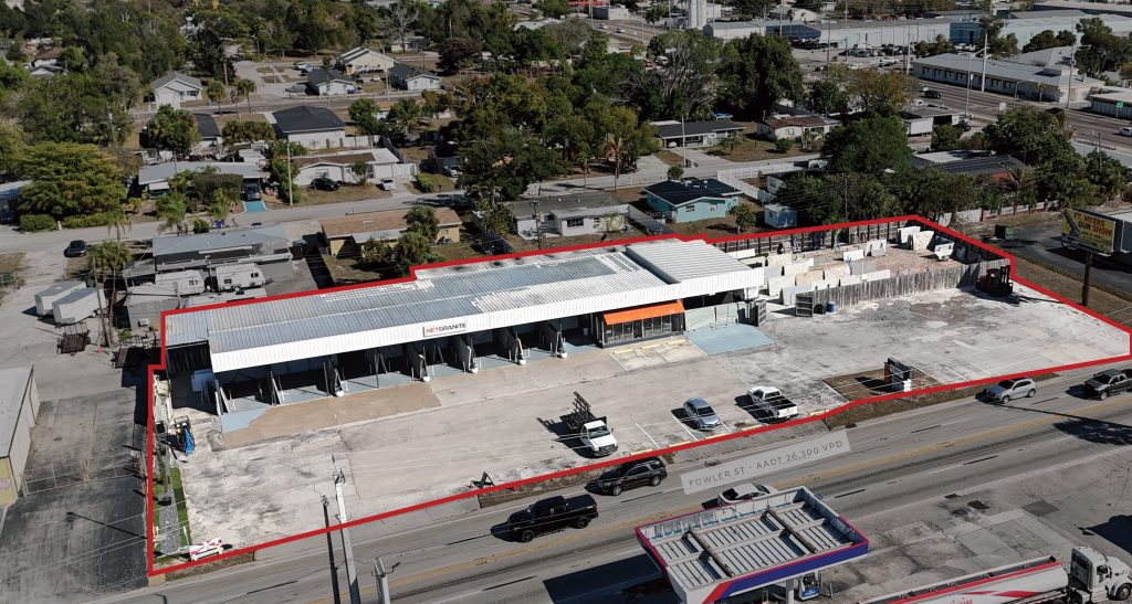 Aerial view of a commercial warehouse with a white roof and multiple loading bays, outlined in red, adjacent to a main road with cars and surrounded by residential buildings.