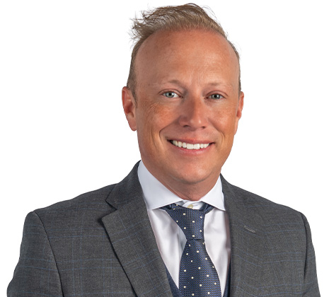 A man wearing a gray plaid suit, white shirt, and blue polka dot tie, smiling at the camera against a white background.