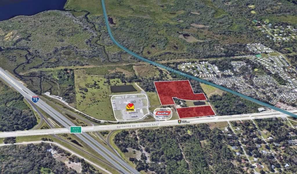 Aerial view of a highway exit featuring a Shell gas station, Rocket car wash, and nearby commercial land plots surrounded by greenery and residential areas.