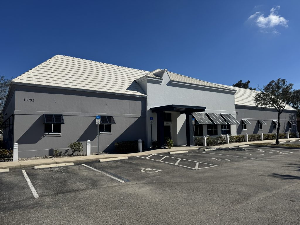 A single-story commercial building with a white roof, gray walls, multiple windows with awnings, and an empty parking lot under a clear blue sky.