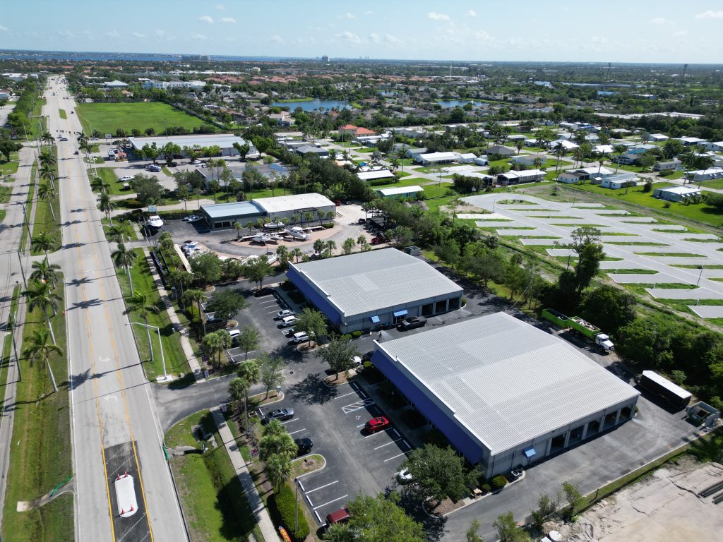 Aerial view of two large industrial buildings with parking lots, a main road on the left, and surrounding green spaces and residential areas in the background.