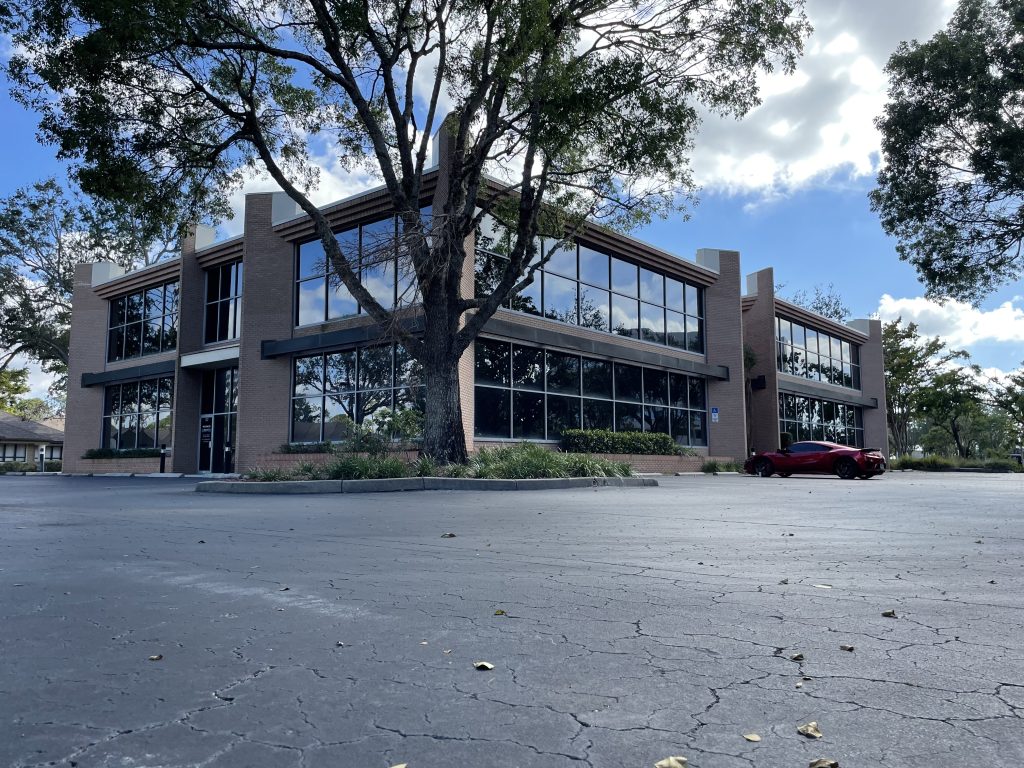 Two-story office building with large glass windows, surrounded by trees and an empty parking lot with a red car parked on the right side.