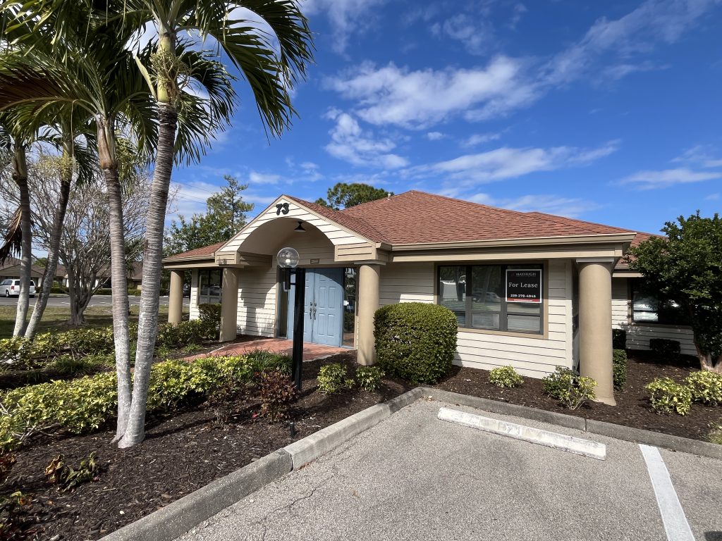 Single-story office building with a red roof, blue double doors, and a "For Lease" sign in the window, surrounded by landscaping and palm trees under a blue sky.