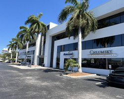 A modern white office building with large windows, palm trees lining the sidewalk, and business signs visible on the ground floor.