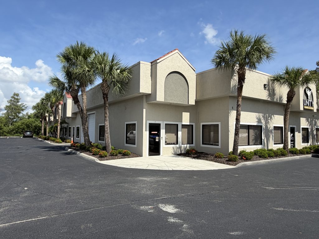 A beige, one-story commercial building with palm trees along the sidewalk, under a blue sky with scattered clouds.