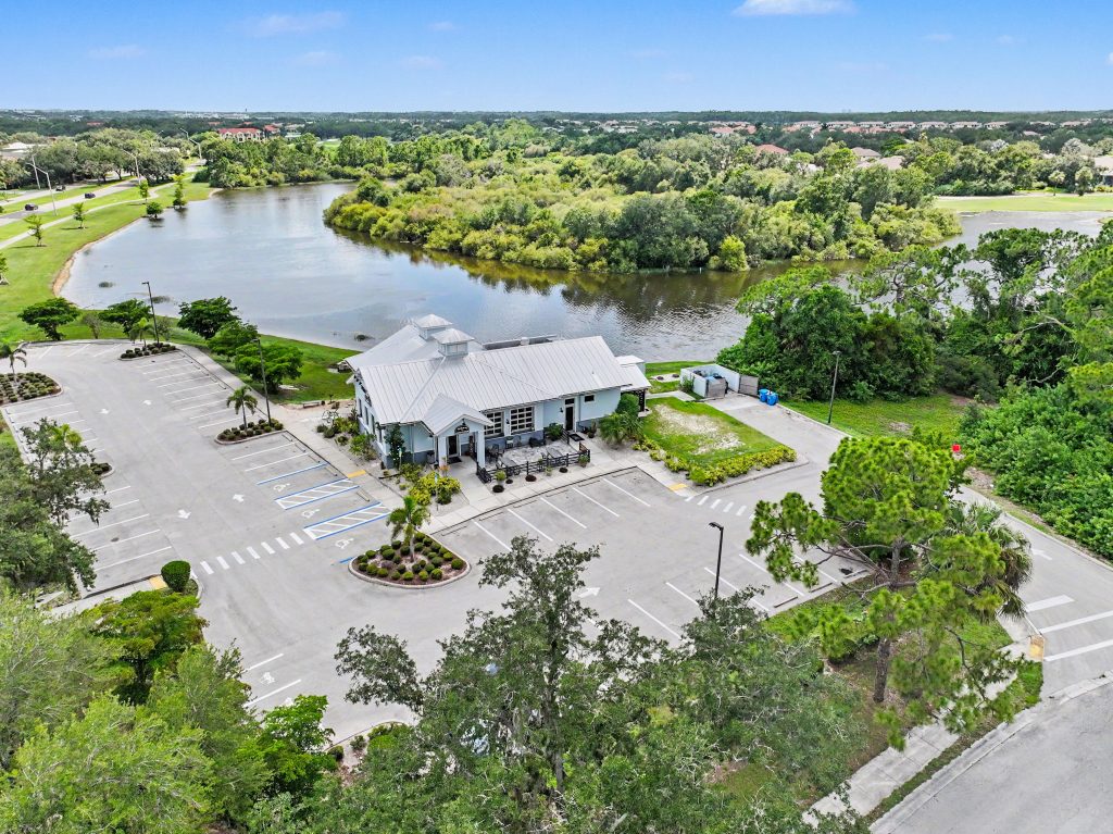 A small building with a metal roof sits beside a lake, surrounded by trees and an empty parking lot under a clear blue sky.