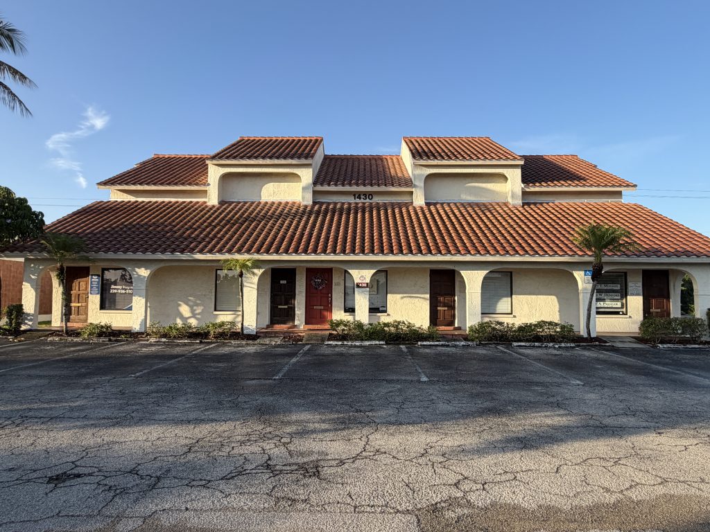 A two-story building with a red tile roof, beige exterior, and multiple doors and windows, set in front of a mostly empty parking lot under a clear sky.