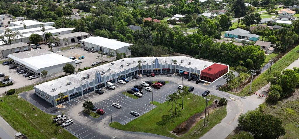 Aerial view of a strip mall with several parked cars; one end storefront is highlighted in red, surrounded by greenery and nearby residential buildings.