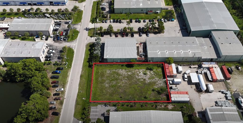 Aerial view of an industrial area with a rectangular, vacant, grassy lot outlined in red, surrounded by warehouses and parked vehicles.