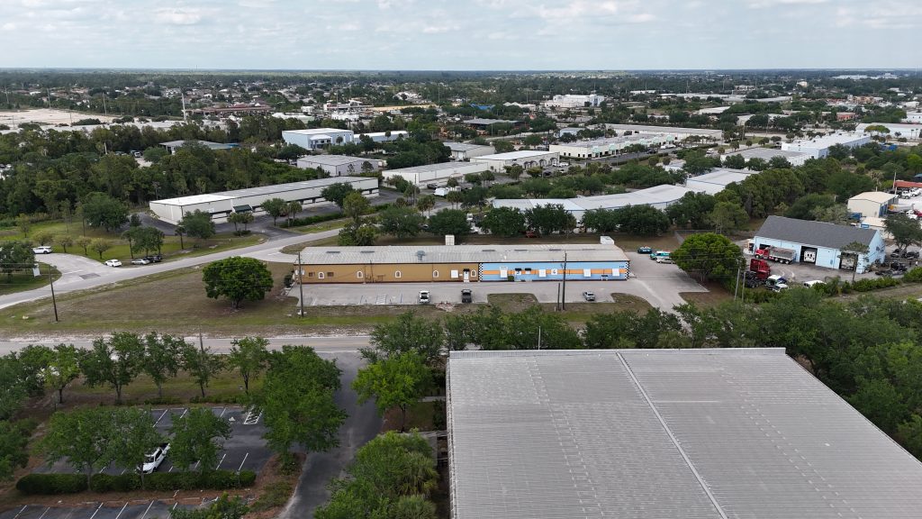 Aerial view of a light industrial area with warehouses, a tan building with blue accents, and scattered trees under a partly cloudy sky.
