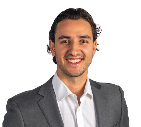 Smiling man with dark hair wearing a white shirt and grey suit jacket, standing against a plain white background.